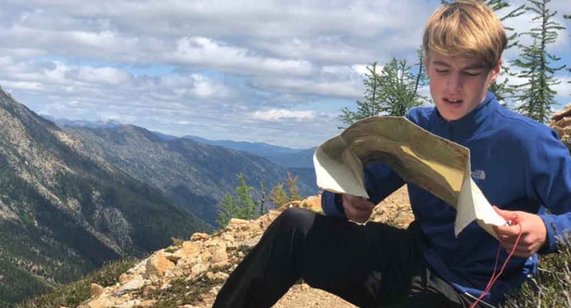 a teen reads a map while sitting on a rock overlooking a mountainous landscape in the pacific northwest